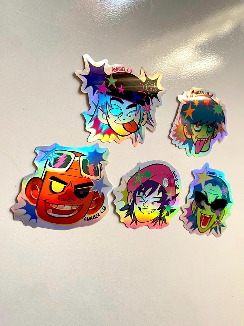 May include: A collection of six holographic stickers featuring cartoon character faces with various expressions. The stickers have a starburst design around the edges and the text "ANABEL CB" is printed on each sticker.