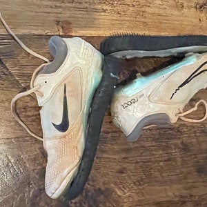 May include: Pair of worn beige and black Nike athletic shoes with black swoosh logos. The shoes are on a wooden surface. The soles are black and show signs of wear. The laces are beige.
