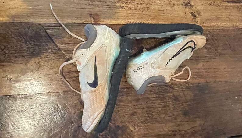 May include: Pair of worn beige and black Nike athletic shoes with black swoosh logos. The shoes are on a wooden surface. The soles are black and show signs of wear. The laces are beige.