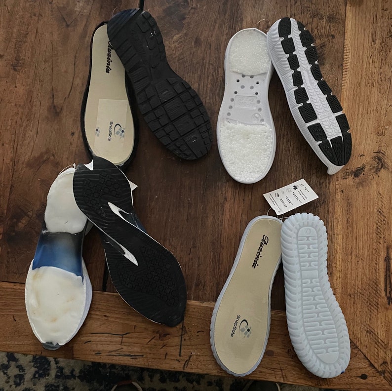 May include: Assorted shoe insoles and outsoles in various colors and designs are arranged on a wooden surface. The insoles feature the brand name "Quzinia" and "SnapSole". The outsoles are black, white, and blue.