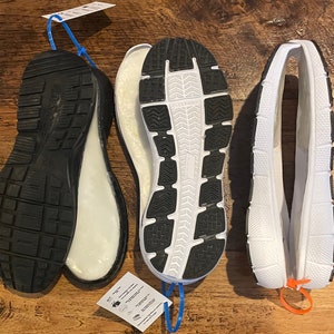 May include: Three shoe soles are displayed, showcasing their design and construction. The soles are black and white, with textured patterns on the bottom for grip. The soles are made of rubber and foam. The image highlights the different components of footwear.