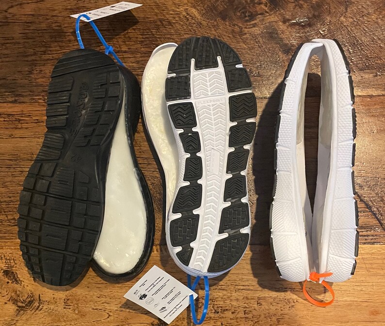 May include: Three shoe soles are displayed, showcasing their design and construction. The soles are black and white, with textured patterns on the bottom for grip. The soles are made of rubber and foam. The image highlights the different components of footwear.