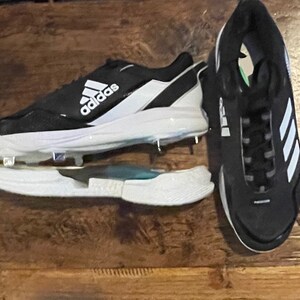 May include: Black and white Adidas baseball cleats with metal spikes. The cleats feature the Adidas logo in white on the side and tongue. The sole is white, and the upper is black with white accents. The cleats are designed for baseball.
