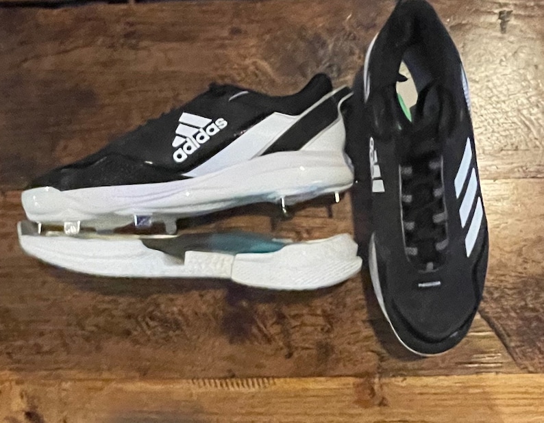 May include: Black and white Adidas baseball cleats with metal spikes. The cleats feature the Adidas logo in white on the side and tongue. The sole is white, and the upper is black with white accents. The cleats are designed for baseball.