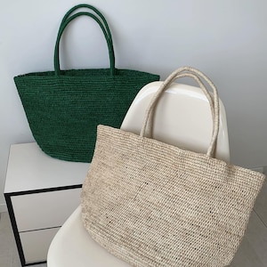 May include: Two woven tote bags. One is a deep green colour, and the other is a natural beige. Both bags have matching woven handles. The bags are sitting on a white chair and a white table.