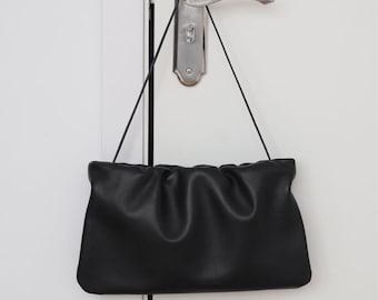 May include: A black clutch purse with a gathered top, hanging from a black strap. The purse is rectangular with a soft, textured appearance. The background is a white door with a silver handle.