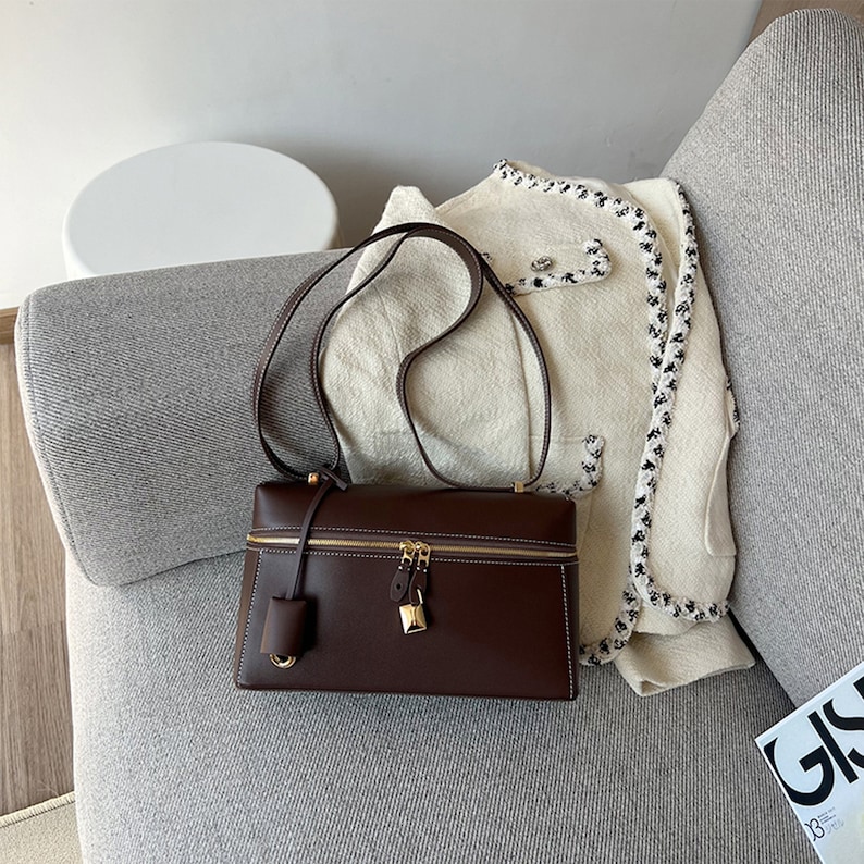 May include: A brown leather satchel bag with a gold zipper and lock. The bag has a long strap and is sitting on a gray couch.