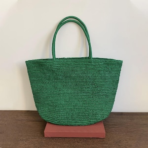 May include: A green woven tote bag with two matching handles. The bag is made of a textured material and has a rounded shape. The bag is sitting on a red rectangular object.