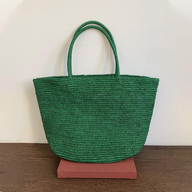 May include: A green woven tote bag with two matching handles. The bag is made of a textured material and has a rounded shape. The bag is sitting on a red rectangular object.