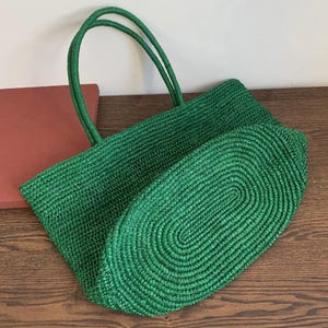 May include: A green woven tote bag with two handles. The bag is made of a textured material and has an oval-shaped base. The handles are a matching green colour. The bag is sitting on a wooden surface.