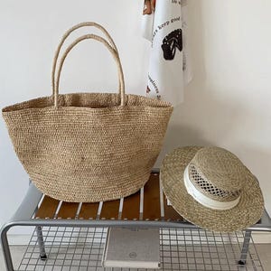 May include: A light brown woven straw tote bag with two handles and a matching straw hat with a white band. The bag and hat are on a metal and wood shelf. The bag is large and open.