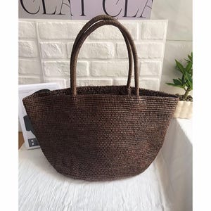 May include: A brown woven tote bag with two sturdy handles. The bag has a rounded shape and a textured surface. The bag is made of a natural material, perfect for carrying items. The bag is sitting on a white surface.