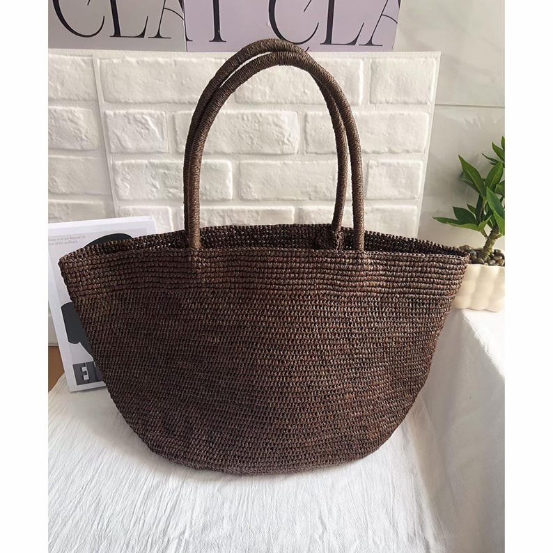 May include: A brown woven tote bag with two sturdy handles. The bag has a rounded shape and a textured surface. The bag is made of a natural material, perfect for carrying items. The bag is sitting on a white surface.
