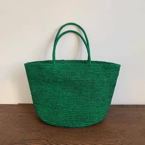 May include: A green woven tote bag with two matching handles. The bag is made of a textured material and has a rounded shape. The bag is on a wooden surface against a white wall.