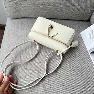 May include: A white leather crossbody bag with a gold zipper and a gold lock closure. The bag has a long adjustable strap.