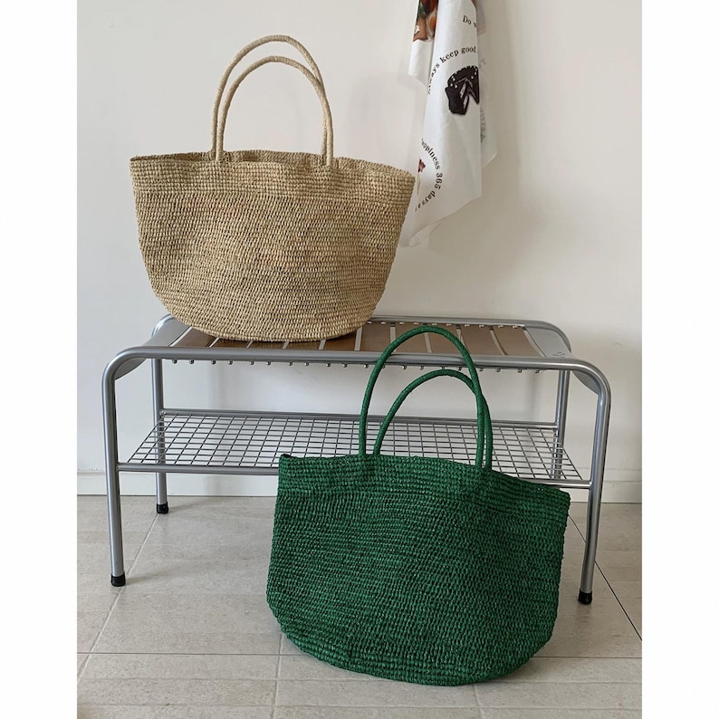 May include: Two woven tote bags, one in a natural tan colour and the other in a deep green. Both bags have matching handles. The bags are on a metal bench with a grid-like shelf.