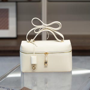 May include: A white leather handbag with a gold zipper and a gold clasp. The bag has a top handle and a long strap that can be worn over the shoulder.