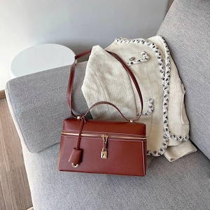 May include: A brown leather satchel handbag with a gold zipper and lock. The bag has a top handle and a long shoulder strap. The bag is sitting on a white and gray couch.