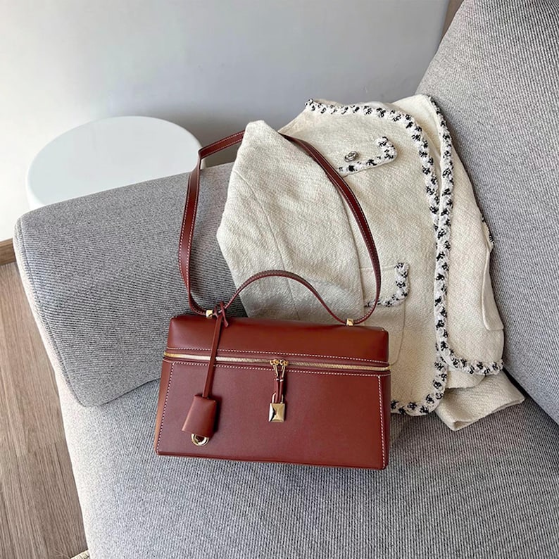 May include: A brown leather satchel handbag with a gold zipper and lock. The bag has a top handle and a long shoulder strap. The bag is sitting on a white and gray couch.