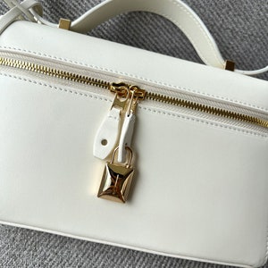May include: A white leather handbag with a gold zipper and a gold padlock closure.