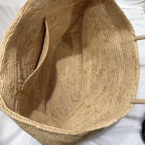 May include: A large, open-top tote bag made of woven natural fibres. The bag has a rectangular shape with a single interior pocket and two handles. The colour is a light tan, with a textured appearance.