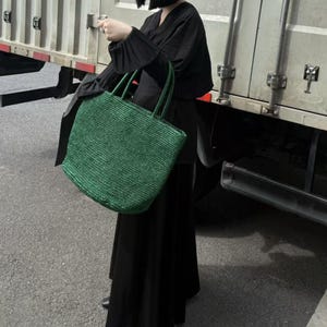 May include: A green woven tote bag with matching handles. The bag is held by a person wearing a black outfit. The bag has a rectangular shape and a textured appearance, suggesting a natural fibre material.