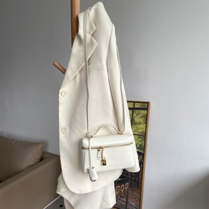 May include: A white leather satchel bag with a gold zipper and lock. The bag is hanging on a white blazer.