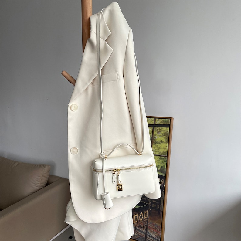 May include: A white leather satchel bag with a gold zipper and lock. The bag is hanging on a white blazer.