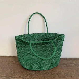 May include: A green woven tote bag with two handles. The bag is made of a textured material and has a rounded shape. The handles are also green and appear to be made of the same material. The bag is sitting on a wooden surface.