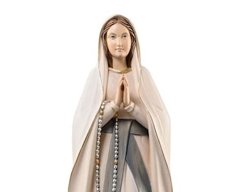 Op de afbeelding: Een wit houten beeld van de Maagd Maria, staand op een bruine rotsbasis met het woord "Lourdes" erop. Ze draagt een witte jurk met een blauwe sjerp en een gouden rozenkrans.