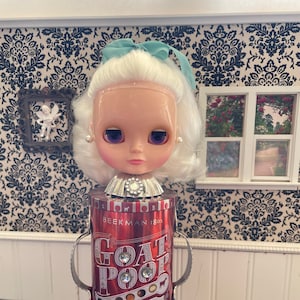 May include: A white doll with blue eyes and a light blue bow in her hair is standing on a red can with the words "Goat Poop" printed on it. The can is decorated with rhinestones and has a silver lid. The can is labelled "Belgian Chocolate Covered" and "with artisan fillings".