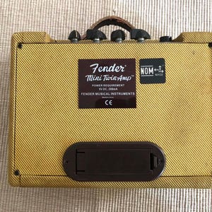 Puede incluir: Un amplificador de guitarra Fender Mini Twin-Amp con un exterior de tweed amarillo. El amplificador tiene un asa de cuero marr&oacute;n, perillas de control negras y un compartimento de bater&iacute;a rectangular marr&oacute;n. Una etiqueta marr&oacute;n dice "Fender Mini Twin-Amp".