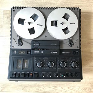 Puede incluir: Un magnetófono de bobina abierta Philips N 4504 en acabado gris y negro. El dispositivo cuenta con dos bobinas plateadas, una unidad 3 motors heads, medidores de nivel analógicos y varios mandos e interruptores de control.