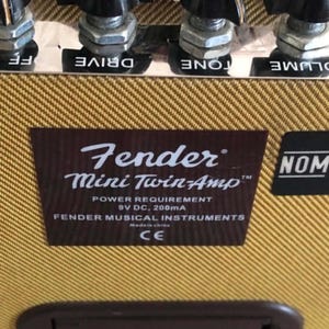 Puede incluir: Primer plano de un amplificador de guitarra Fender Mini Twin-Amp. El amplificador tiene una cubierta de tweed marr&oacute;n y beige con perillas de control negras. La etiqueta del amplificador dice "Fender Mini Twin-Amp" e incluye los requisitos de energ&iacute;a. Tambi&eacute;n es visible una pegatina negra con "NOM".