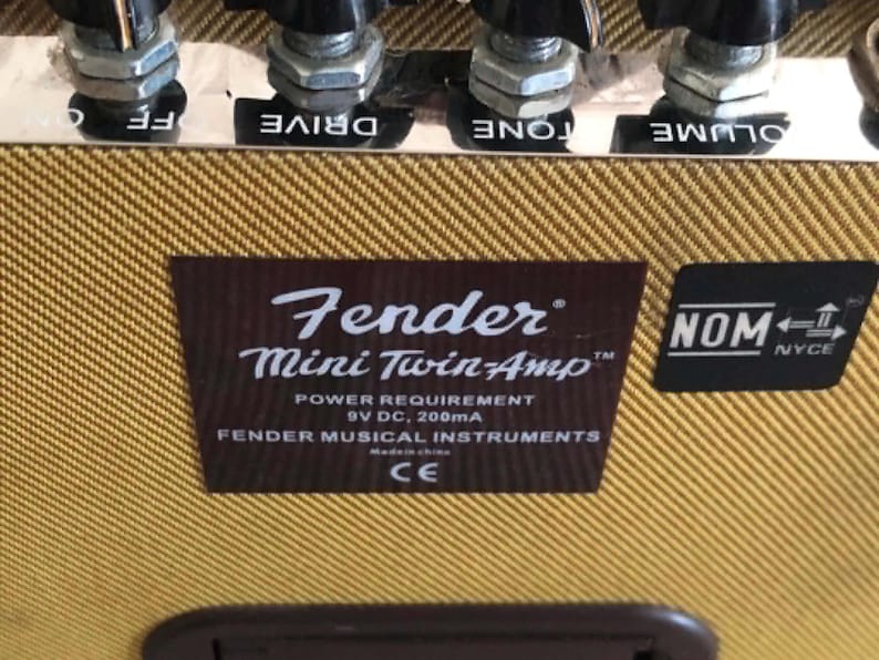 Puede incluir: Primer plano de un amplificador de guitarra Fender Mini Twin-Amp. El amplificador tiene una cubierta de tweed marr&oacute;n y beige con perillas de control negras. La etiqueta del amplificador dice "Fender Mini Twin-Amp" e incluye los requisitos de energ&iacute;a. Tambi&eacute;n es visible una pegatina negra con "NOM".