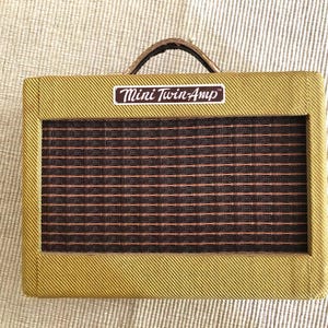 Puede incluir: Un peque&ntilde;o amplificador rectangular con un exterior estilo tweed. La parte frontal presenta una rejilla de altavoz con un patr&oacute;n de cuadr&iacute;cula marr&oacute;n y rojo. La parte superior tiene un asa marr&oacute;n y una etiqueta que dice "Mini Twin-Amp".