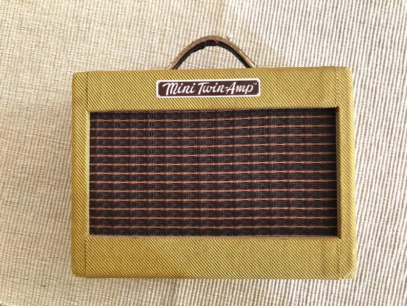 Puede incluir: Un peque&ntilde;o amplificador rectangular con un exterior estilo tweed. La parte frontal presenta una rejilla de altavoz con un patr&oacute;n de cuadr&iacute;cula marr&oacute;n y rojo. La parte superior tiene un asa marr&oacute;n y una etiqueta que dice "Mini Twin-Amp".
