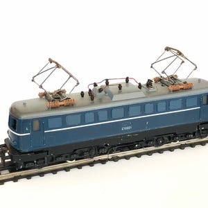 May include: A model electric locomotive, predominantly blue and grey, with detailed overhead pantographs. The miniature train is displayed on a section of railway track, highlighting its realistic construction.
