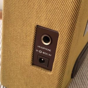 Puede incluir: Primer plano de un peque&ntilde;o amplificador de color beige con un panel marr&oacute;n. El panel tiene una toma de auriculares y una entrada de alimentaci&oacute;n de 9 V CC. El amplificador tiene un exterior de tela texturizada.