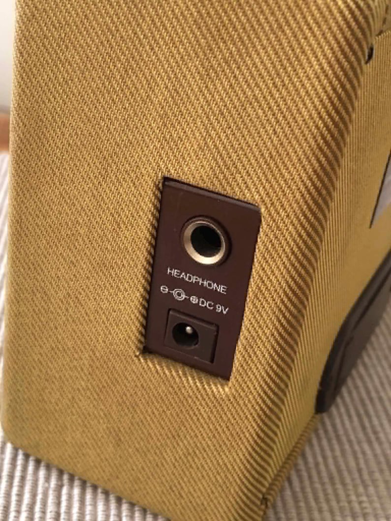 Puede incluir: Primer plano de un peque&ntilde;o amplificador de color beige con un panel marr&oacute;n. El panel tiene una toma de auriculares y una entrada de alimentaci&oacute;n de 9 V CC. El amplificador tiene un exterior de tela texturizada.