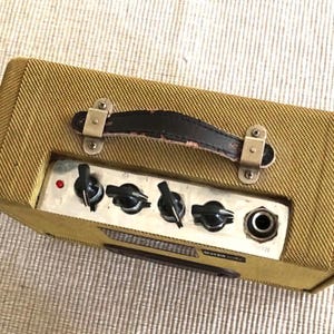 Puede incluir: Un peque&ntilde;o amplificador de guitarra rectangular con un exterior de tela de tweed. Cuenta con un asa de cuero negro, herrajes met&aacute;licos y un panel de control con perillas y un conector. El amplificador es de color marr&oacute;n dorado.
