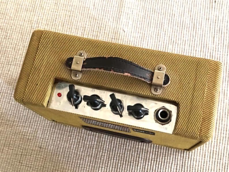 Puede incluir: Un peque&ntilde;o amplificador de guitarra rectangular con un exterior de tela de tweed. Cuenta con un asa de cuero negro, herrajes met&aacute;licos y un panel de control con perillas y un conector. El amplificador es de color marr&oacute;n dorado.
