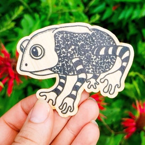 May include: A wooden cutout of a black and white frog with a speckled pattern. The frog has a large eye and is holding its legs up in the air.