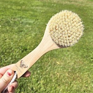 May include: A round body brush with light-colored natural bristles and a smooth wooden handle. The handle features a small engraved logo of a dog and the text Liberty Ranch Company. A white cotton rope loop is attached to the end of the handle.