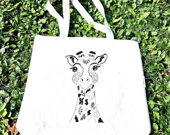 May include: A white canvas tote bag with a black and white giraffe illustration. The giraffe has detailed patterns on its face and neck. The bag has two white handles and is sitting on green grass.