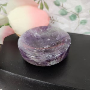 May include: A purple amethyst crystal dish with a smooth, polished surface. The dish is round and has a slight indentation in the center.