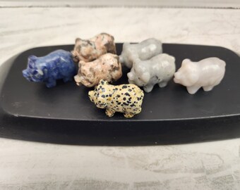 May include: Seven small pig figurines carved from different types of stone. The pigs are in various colors, including blue, white, pink, and brown with black spots.