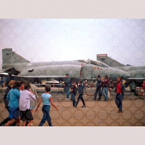 May include: Vintage photograph of two military fighter jets on display, with a crowd of people in the foreground. The jets are painted in a camouflage pattern. The image has a faded, aged appearance, suggesting it was taken some time ago.