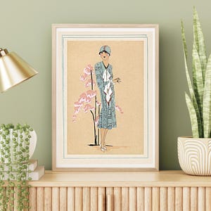 1920s Art Deco fashion illustration from Tres Parisien magazine showing a stylish woman in powder blue dress with white tie collar and matching cloche hat, standing beside pink cherry blossoms. Digital wall art from French vintage fashion publication.