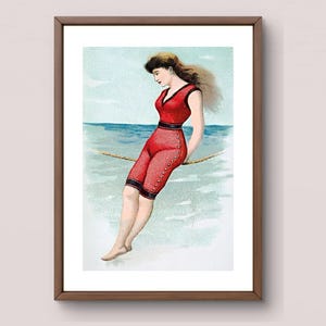 May include: Vintage framed print of a woman in a red swimsuit, seated on a rope over water. The artwork features a woman with brown hair, wearing a one-piece swimsuit with black trim and buttons. The background shows a light blue sky and water.